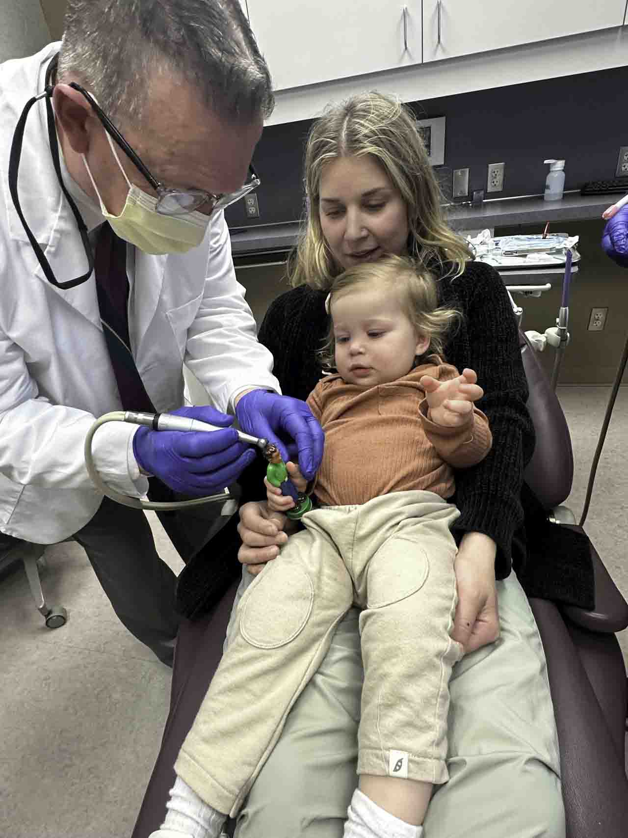 Dr. Kapp showing dental tools to a young child sitting on her mother’s lap in the examination chair.